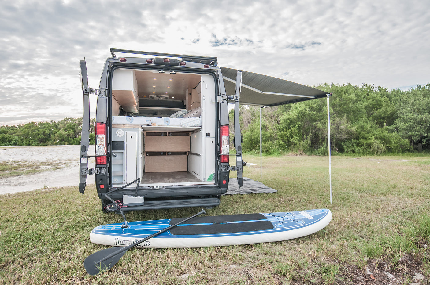 This rv rental camper van shows its cozy interior, extended awning, and paddleboard on the grass, ready for adventure.