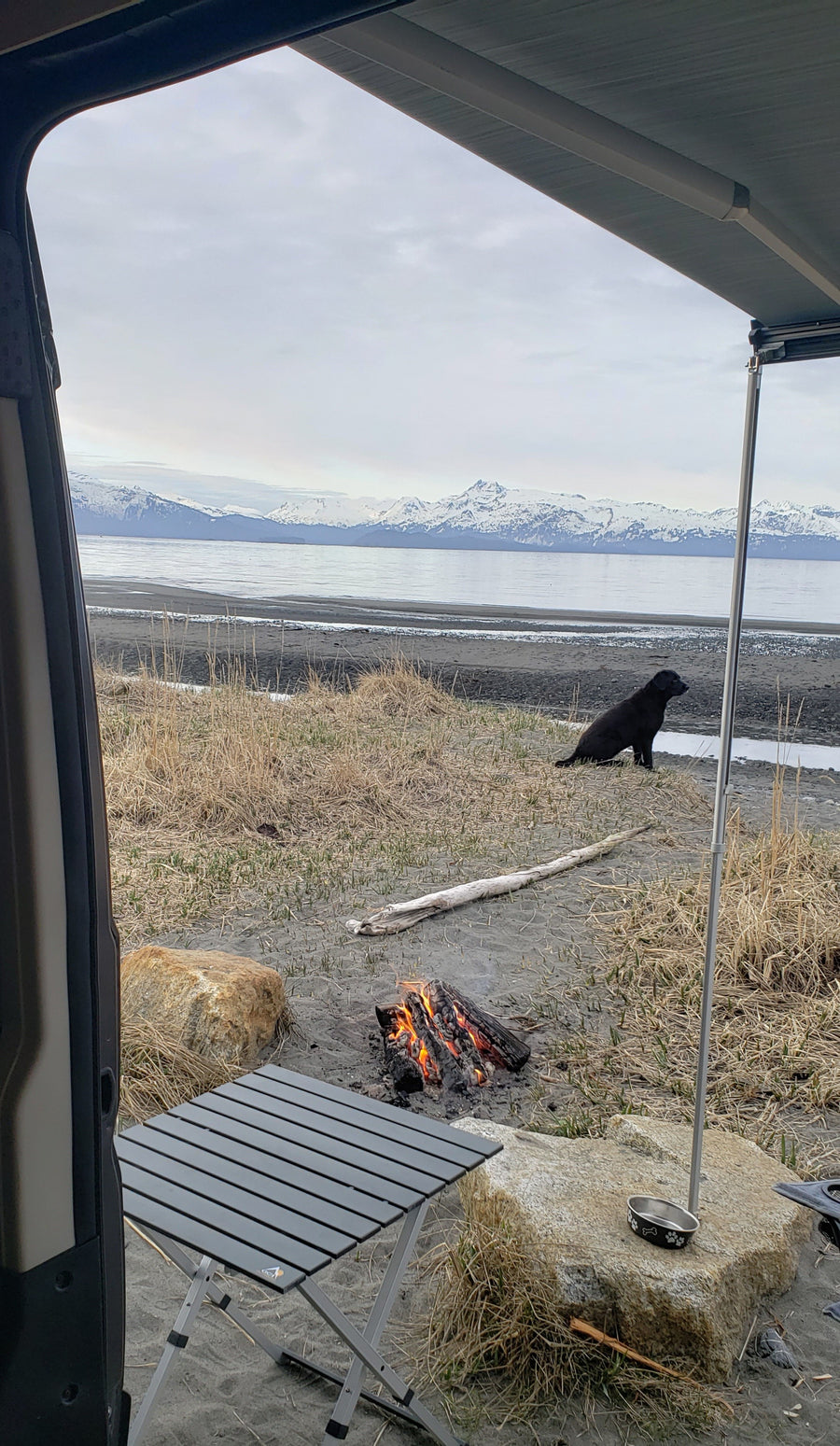 Scenic RV rental view: dog, campfire, and table on a beach with snow-capped mountains across the water.