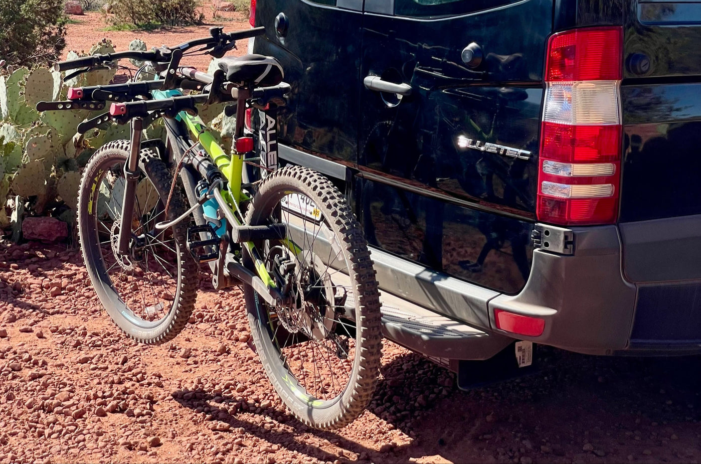 Two mountain bikes are secured on an RV rental van's rear rack, parked on red desert ground, ready for adventure.
