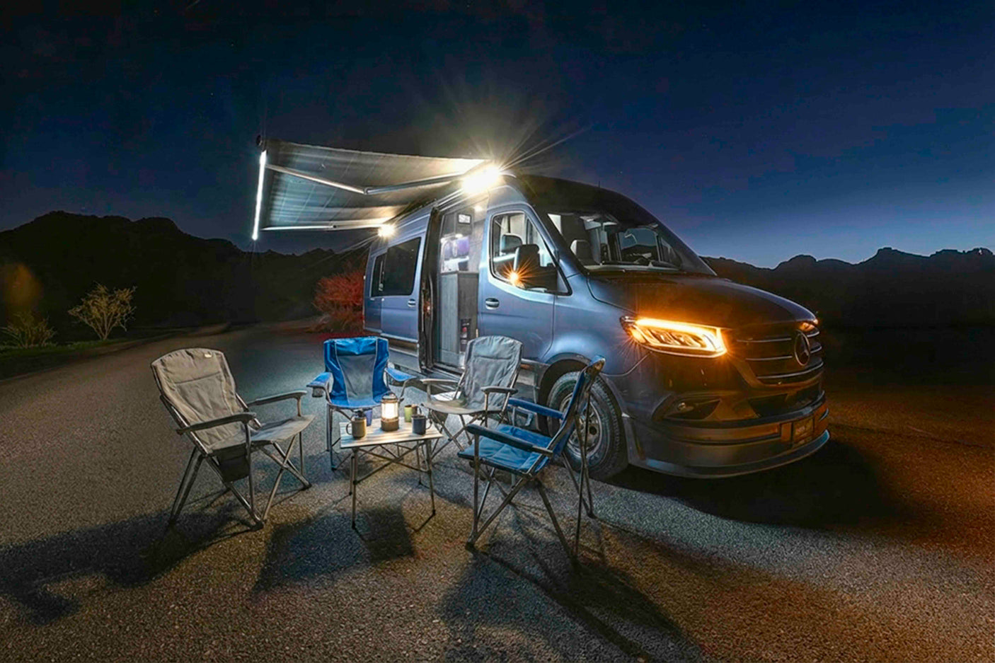 RV rental campout at night: a blue camper van with bright awning lights, headlights, camp chairs, and a lantern-lit table.