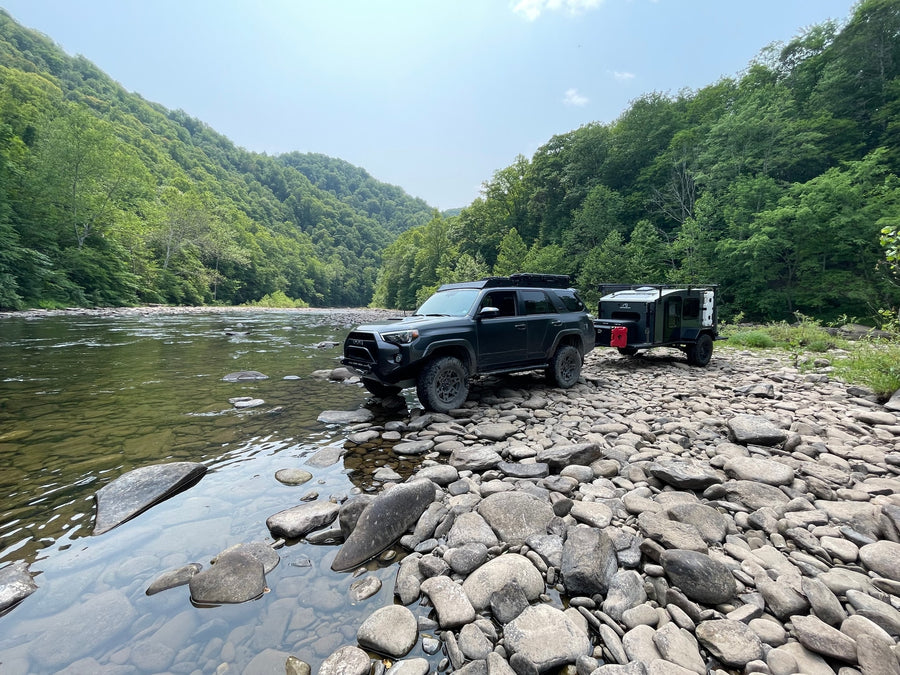 Dark SUV & off-road RV rental parked on a rocky riverbank. Lush green mountains line a clear river