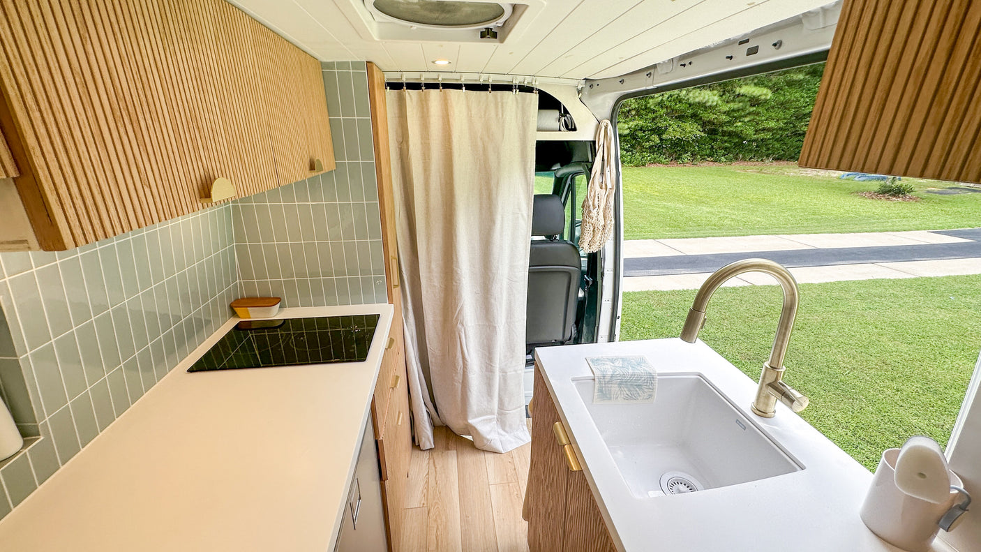 Step into this chic RV rental! Features a compact kitchen with a sink, cooktop, wood cabinets, & outdoor views.
