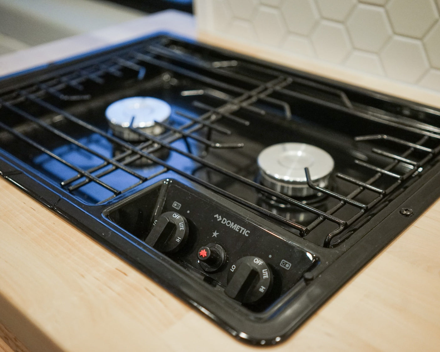 A Dometic two-burner gas cooktop in an RV rental, with one burner lit showing a blue flame, ideal for camp cooking.