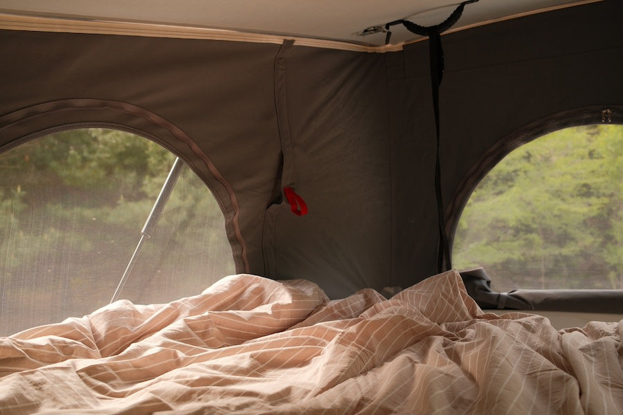Pop-top RV rental interior: A cozy bed with striped linens overlooks a forest through screened windows. Enjoy nature.