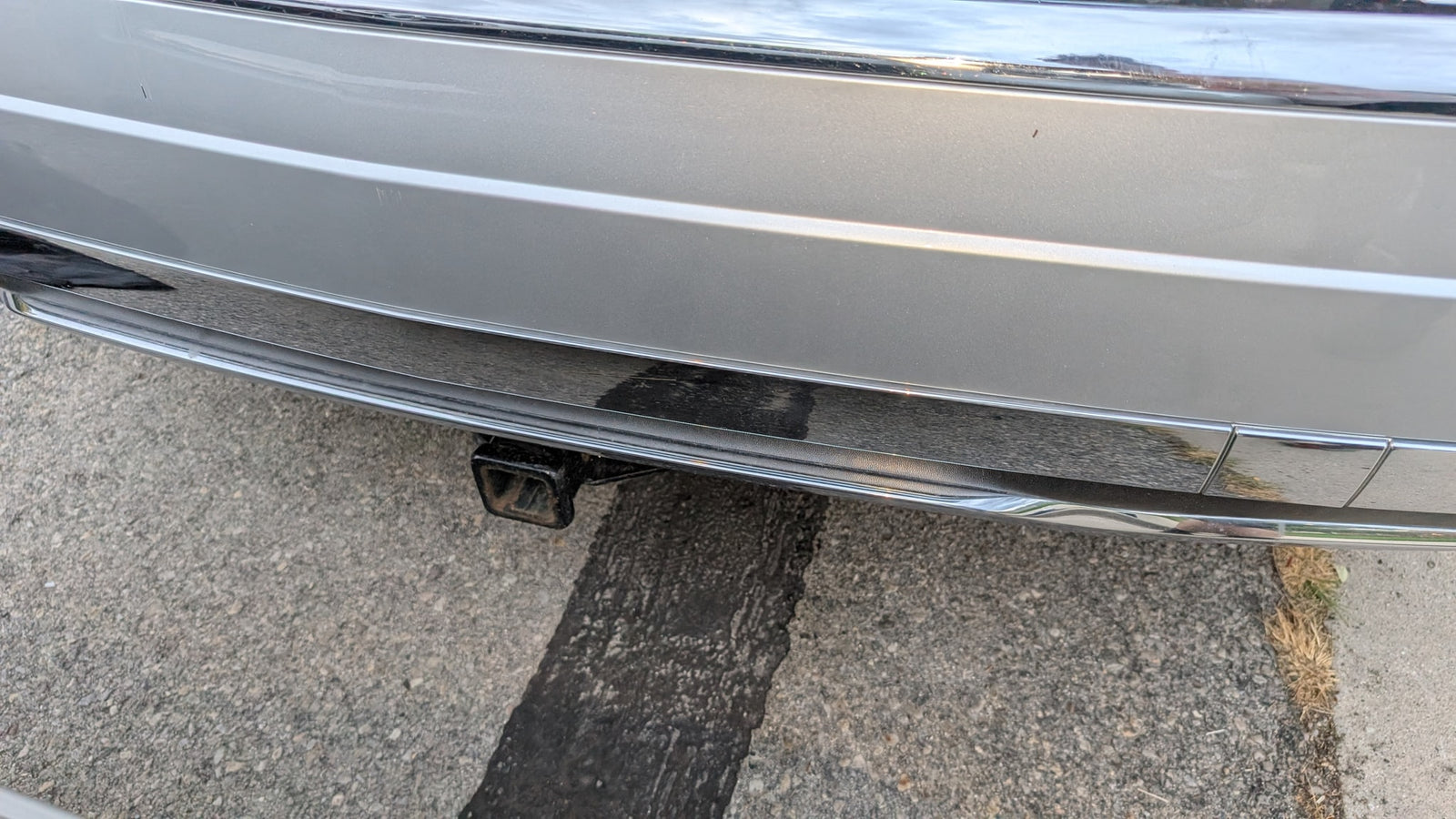 Silver RV rental rear bumper with chrome trim, minor scratches, and a trailer hitch, parked on asphalt.