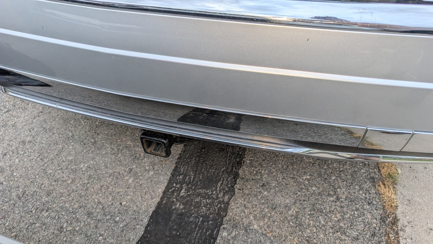 Silver RV rental rear bumper with chrome trim, minor scratches, and a trailer hitch, parked on asphalt.