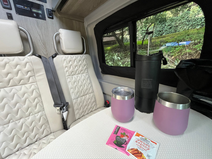 Relaxing RV rental interior: Seats, table with tea/mugs. Solar power setup on the wall. Lush greenery outside window.