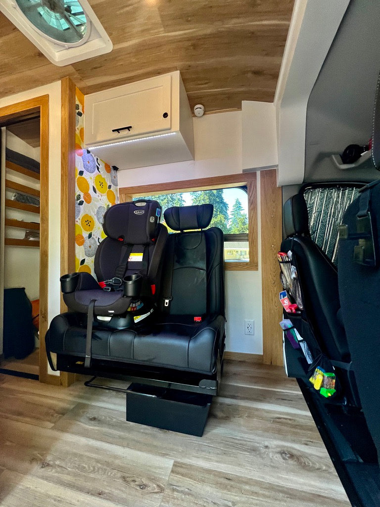 An RV rental's interior shows a child car seat on custom seating, a cabinet, and wooden floors, perfect for family adventures.