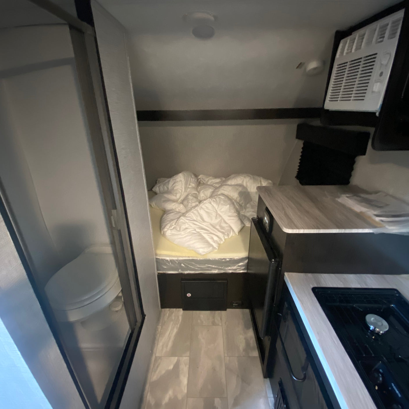Cozy RV rental interior: featuring a compact kitchen, bed with protective wrap, and a shower/toilet bathroom on the left.