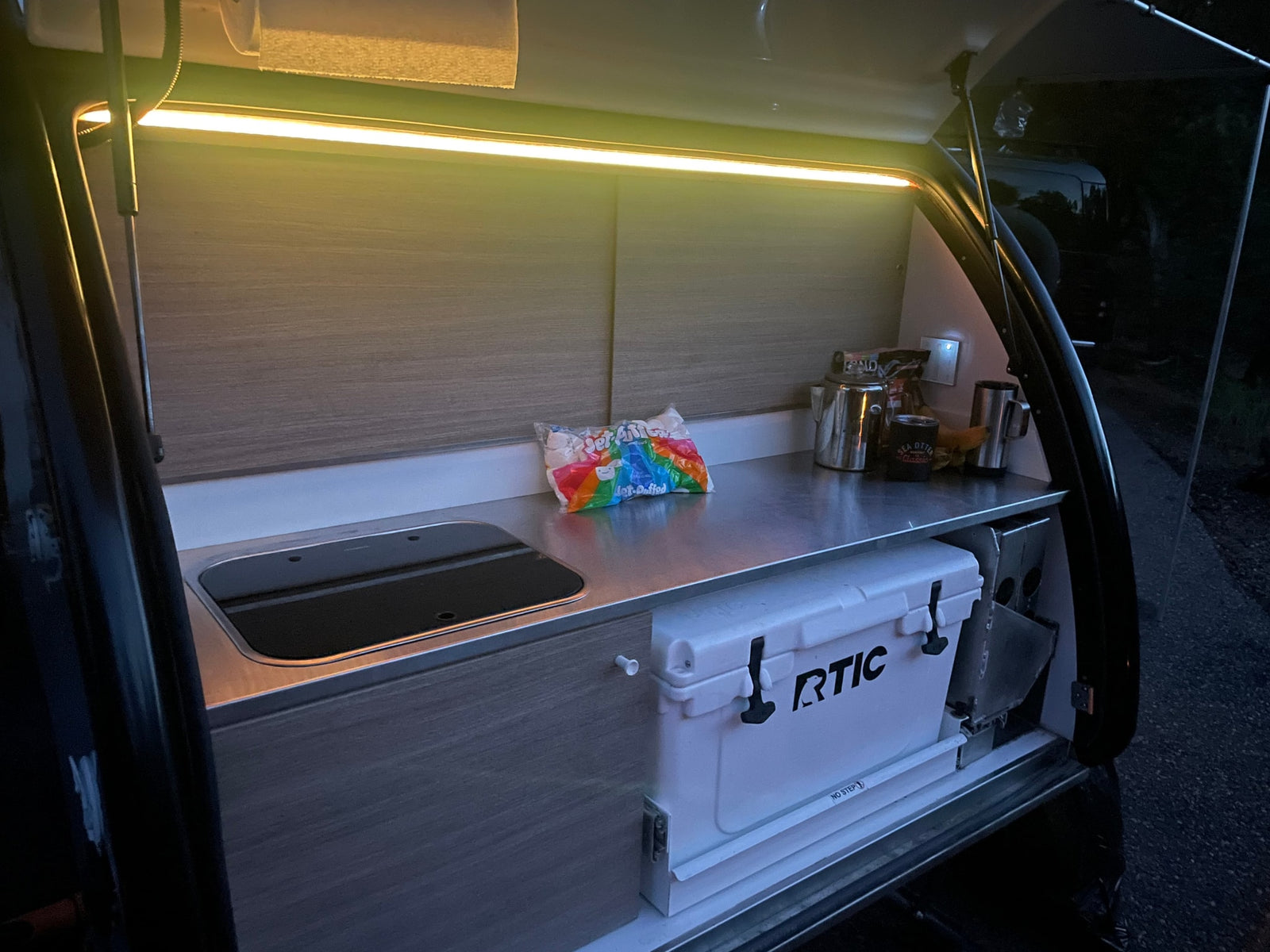 overlooking a scenic forest at sunset.|Black Rivian SUV towing a white teardrop camper on a dirt road. Explore nature with this ideal RV rental setup.|Bright RV rental kitchen/galley: stainless counter