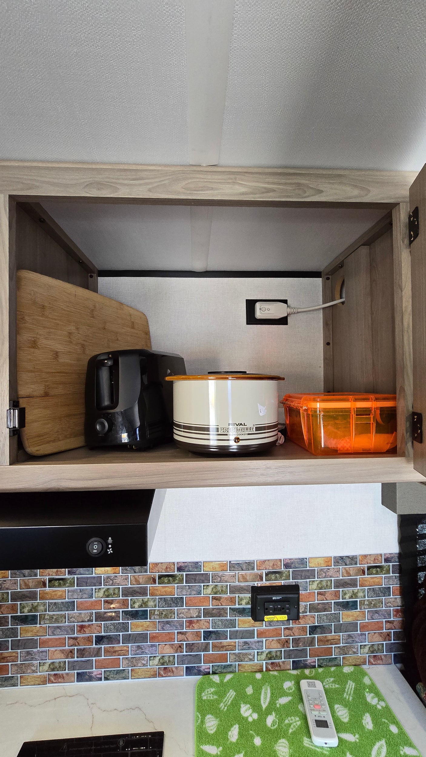 Upper cabinet in an **rv rental** kitchen holds a toaster, slow cooker, and cutting board. Below, a range hood and tiled backsplash.