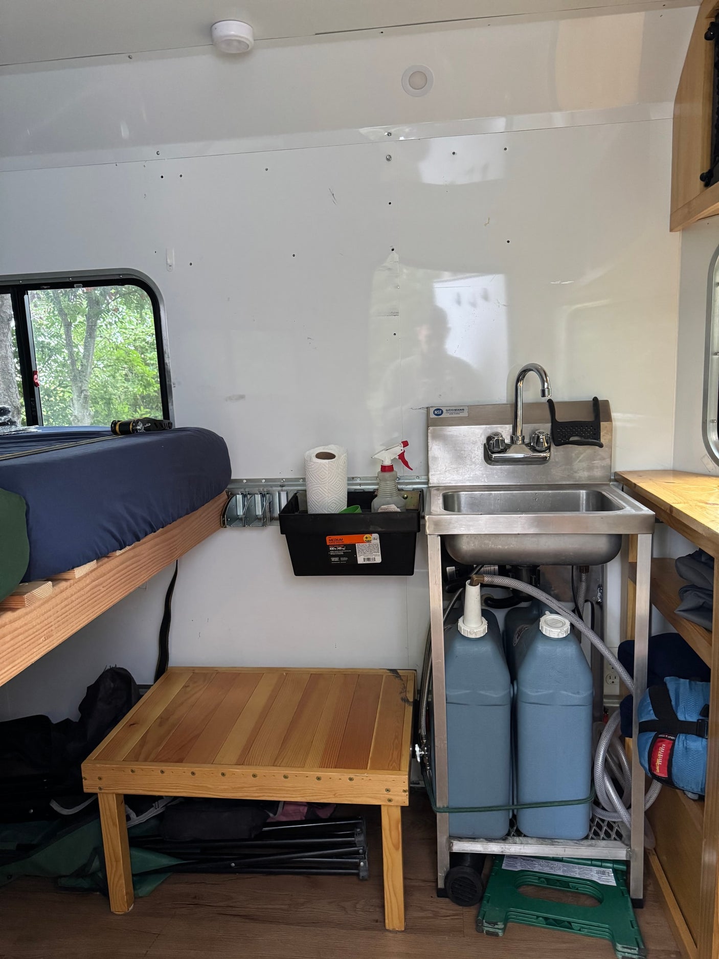Cozy RV rental interior with a bed, wooden table, and compact kitchenette featuring a sink and water storage.