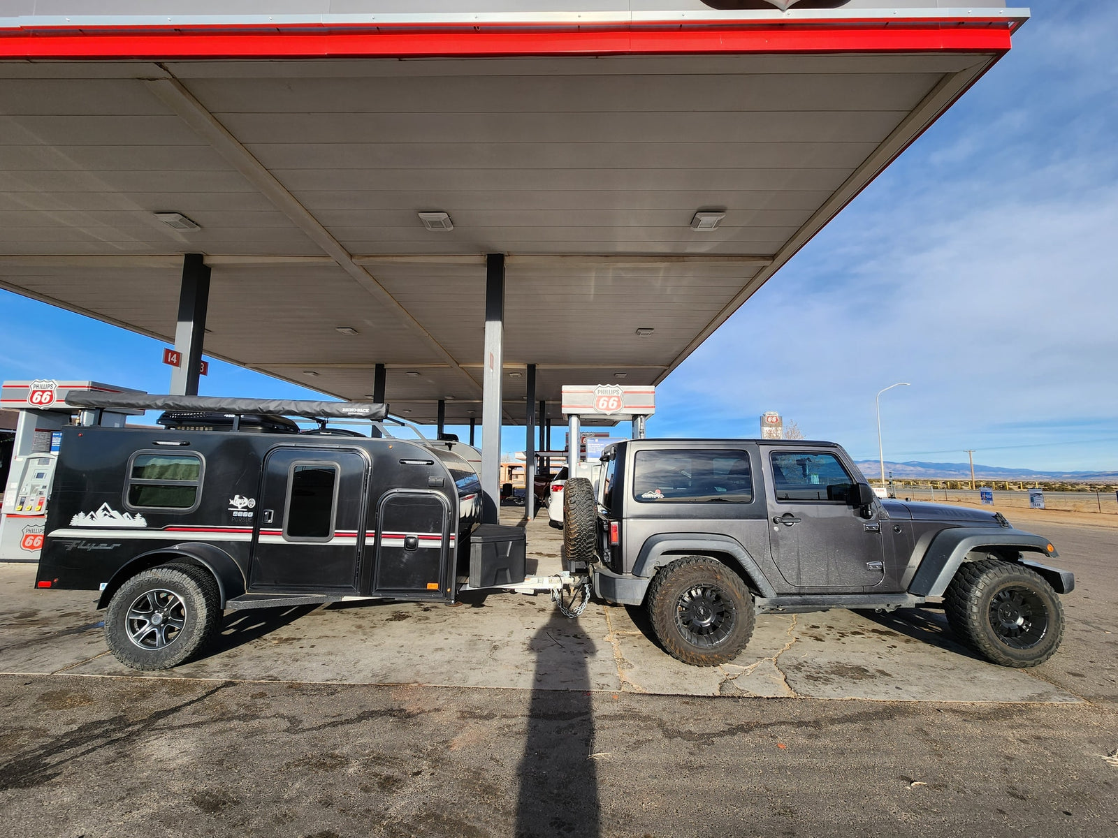 This dark gray Jeep tows a compact RV rental, fueling up at a Phillips 66 station for off-road adventure & camping.