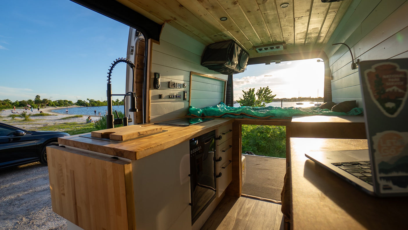Inside an RV rental camper van: kitchenette, bed, and laptop, with a scenic view of a sunny beach and calm water.