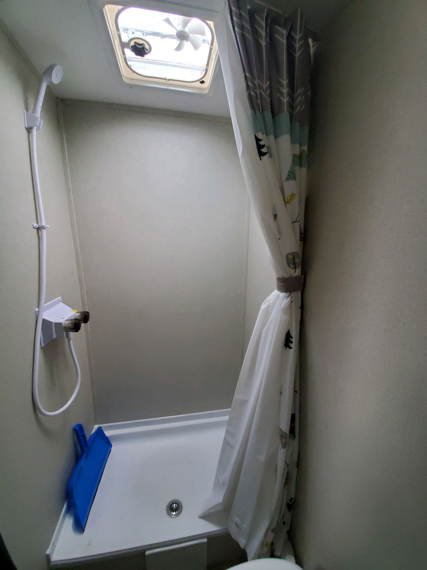 Compact RV rental shower with white pan, handheld showerhead, and a nature-themed curtain. Features a ceiling fan.