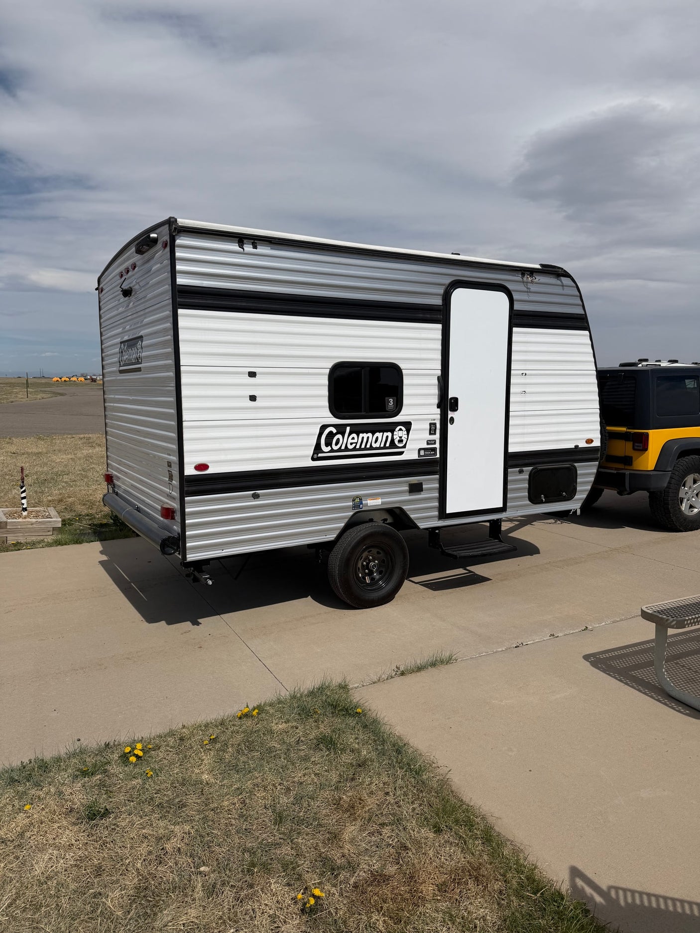 Compact Coleman travel trailer RV rental with a yellow Jeep, parked on concrete under a cloudy sky.