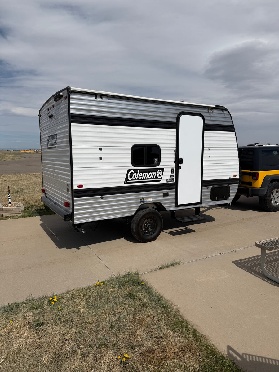 Compact Coleman travel trailer RV rental with a yellow Jeep, parked on concrete under a cloudy sky.
