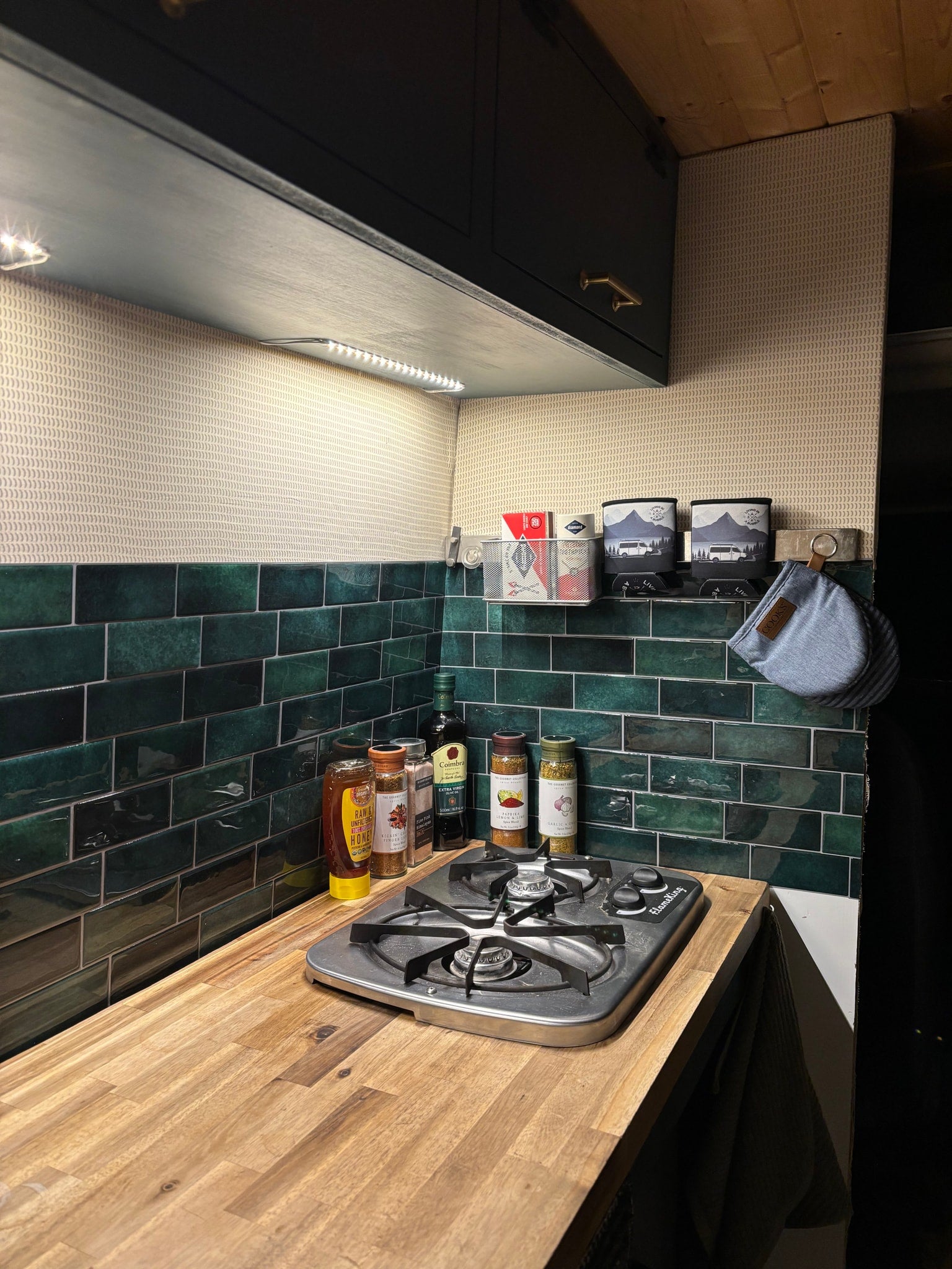 Cozy RV rental kitchen features a compact stove, wooden countertop, green tiled backsplash, and LED lighting for compact living.