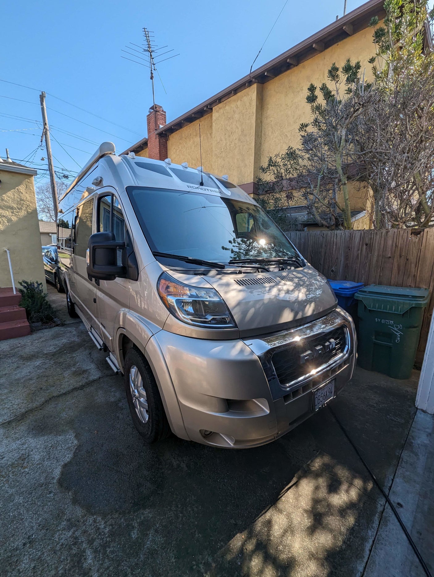 Beige Roadtrek camper van, a perfect RV rental, parked in a sunny driveway by a house and fence.