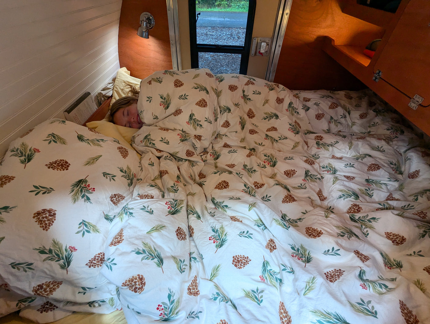 A person smiles peacefully while sleeping cozily in an RV rental bed, covered by festive pinecone and berry bedding.