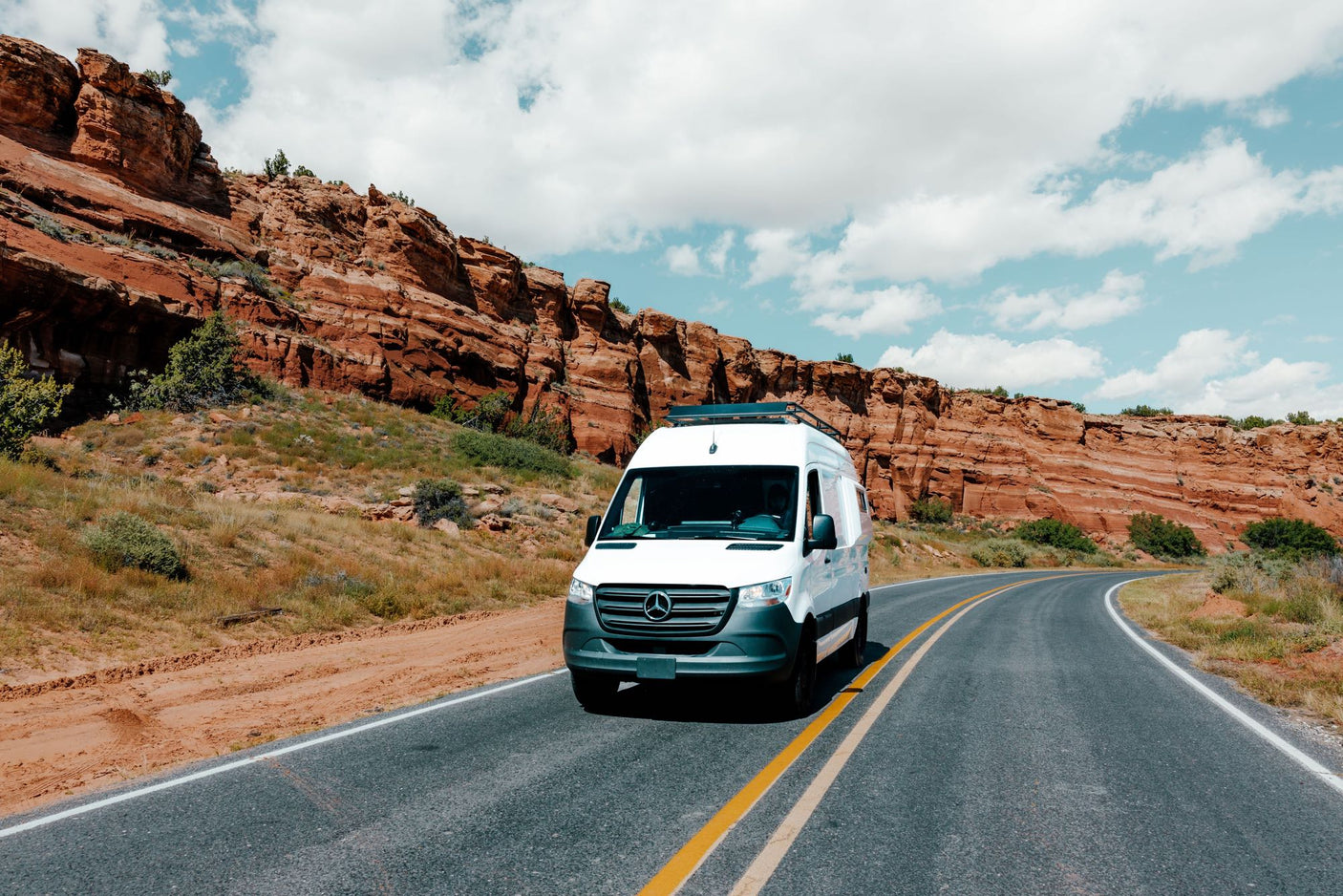 White RV rental van on a desert road, flanked by majestic red rock formations under a partly cloudy sky.