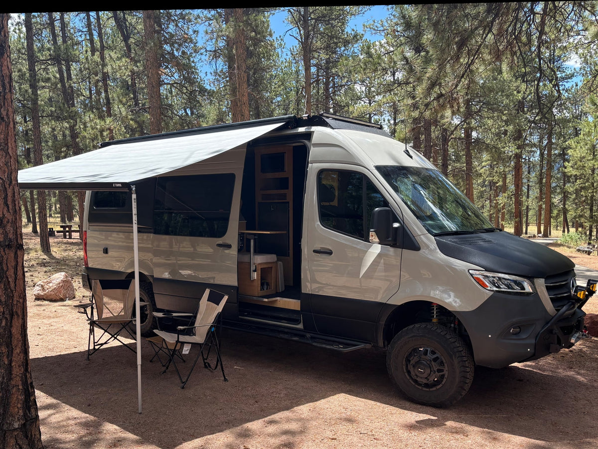 Custom Mercedes-Benz Sprinter camper van rv rental at a pine forest campsite with awning and chairs. Ready for outdoor adventures.|Modern RV rental interior featuring a bamboo kitchen with induction cooktop — photo 1