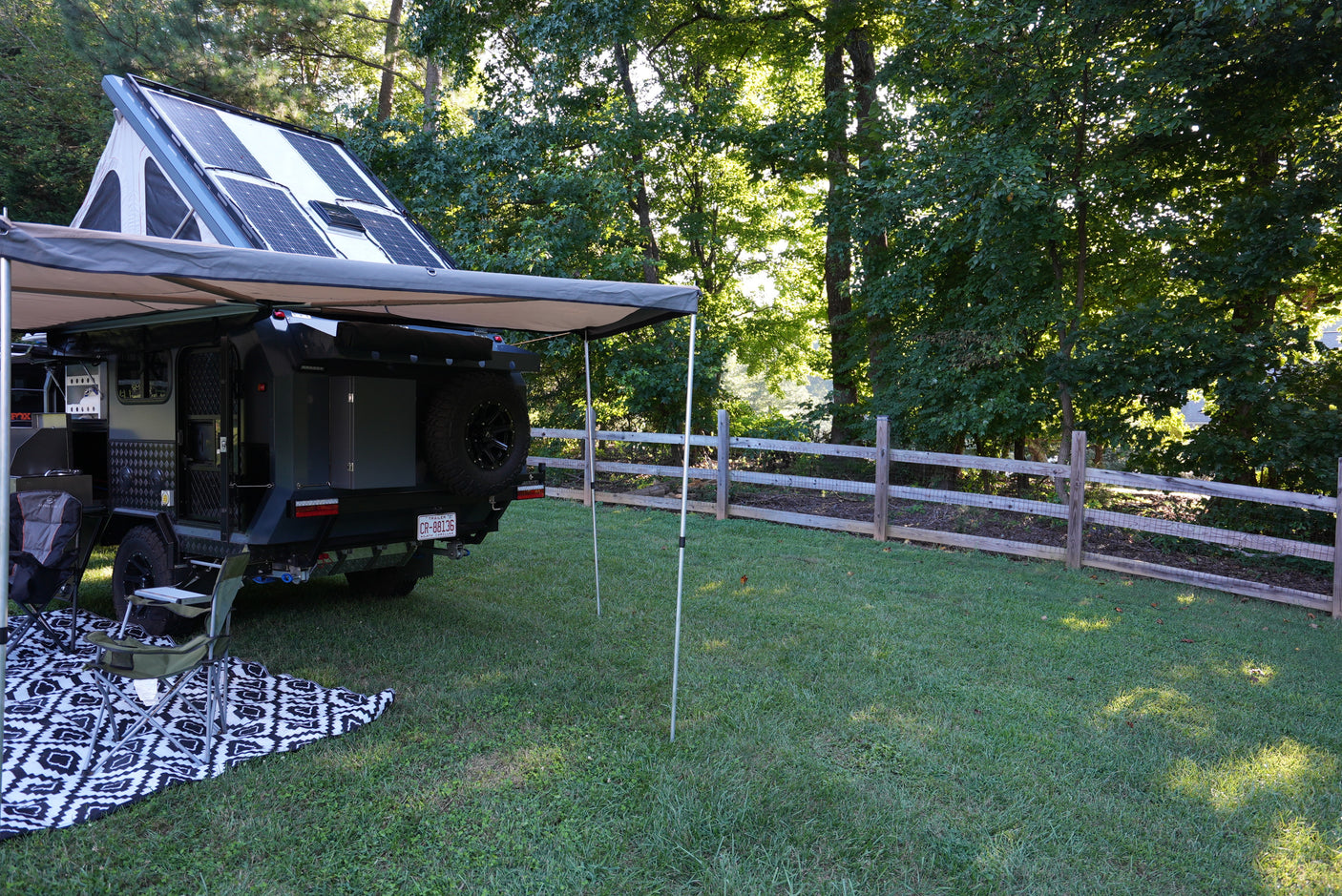 RV rental camper, with rooftop tent, solar panels, & awning, set up in a grassy yard next to trees and a fence.