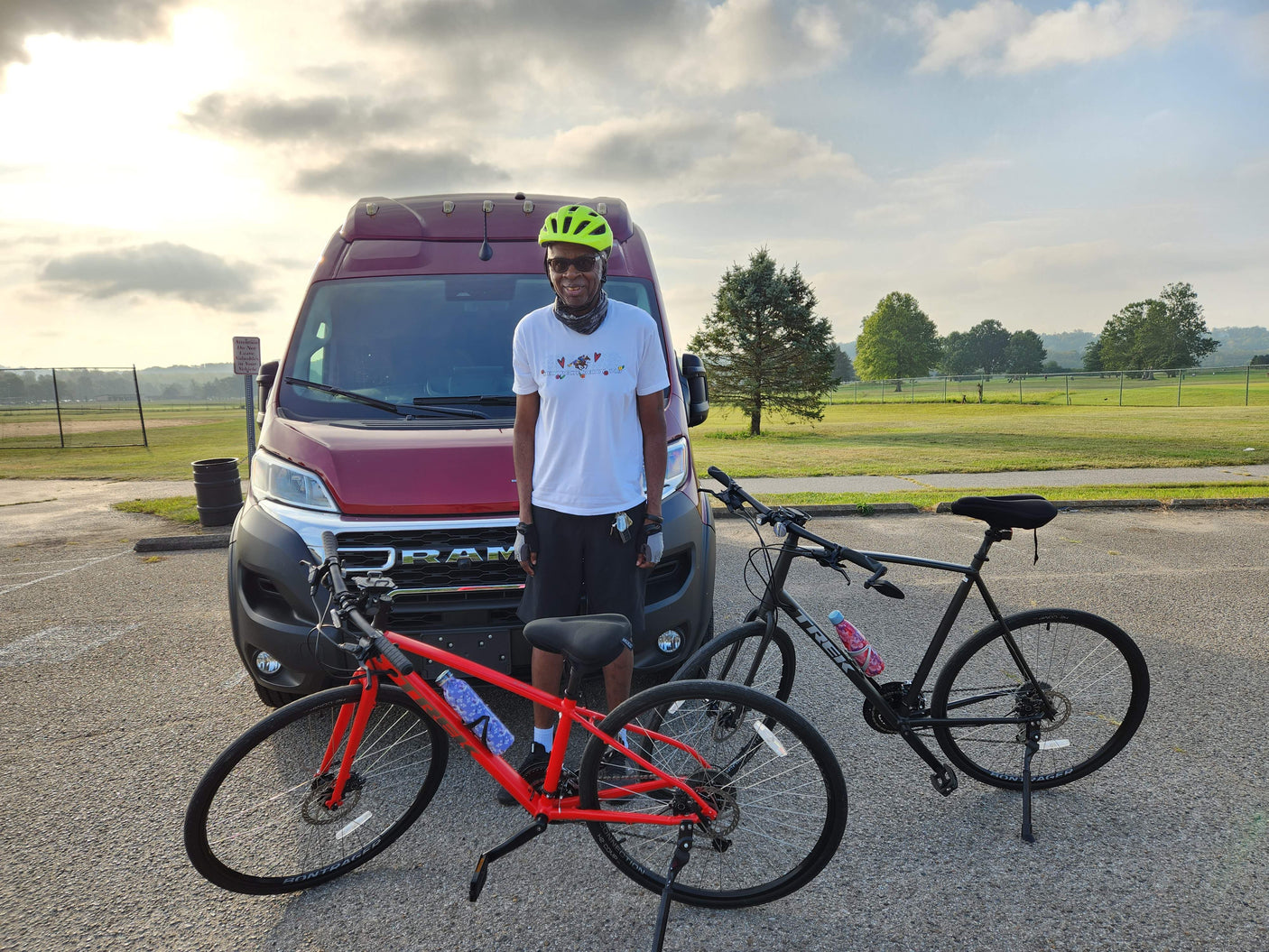 A man in a yellow helmet and white shirt stands with two bikes (red & black) by a red RV rental van in a park.