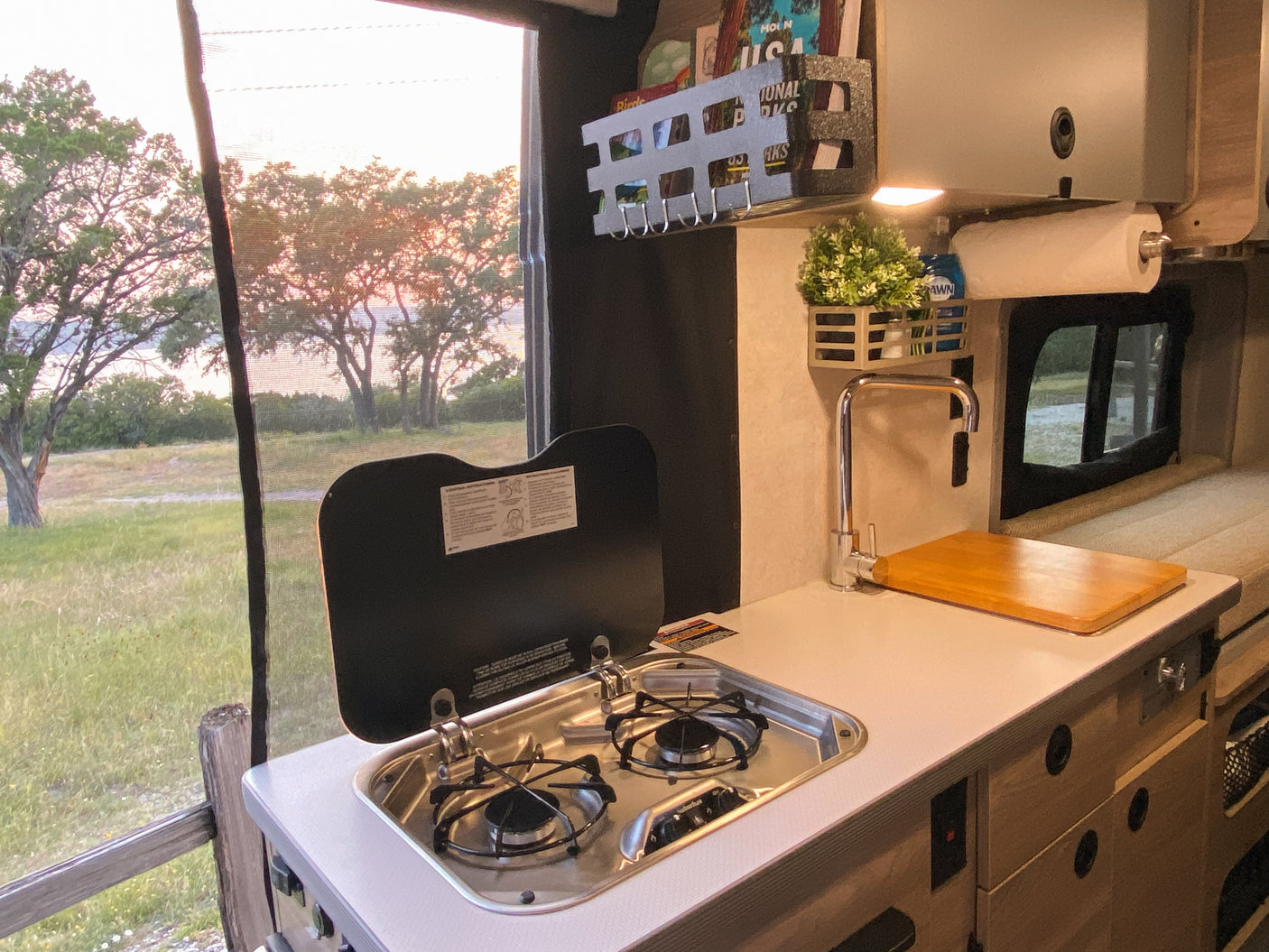 RV rental: Bright camper kitchenette with stove, sink, and scenic window views of trees, grass, and distant water.