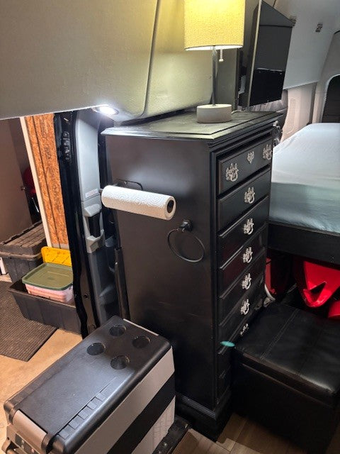 This RV rental features efficient van life design: a tall dresser with a lamp, TV, cooler, and ottoman provides compact storage.