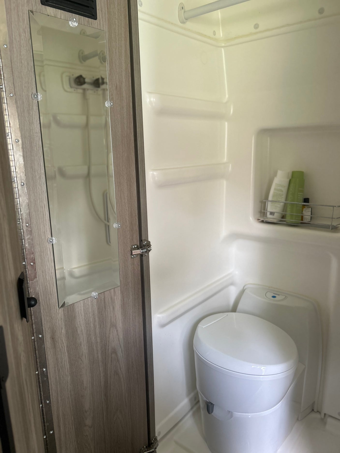 An RV rental's compact wet bath showing a white toilet, shower area with toiletries, and a mirror on the wooden door.