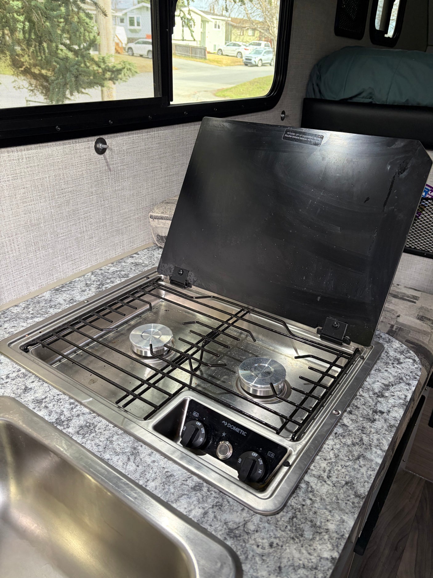 RV rental kitchen: Dometic two-burner stove, sink, and countertop, with a window view of trees and houses.