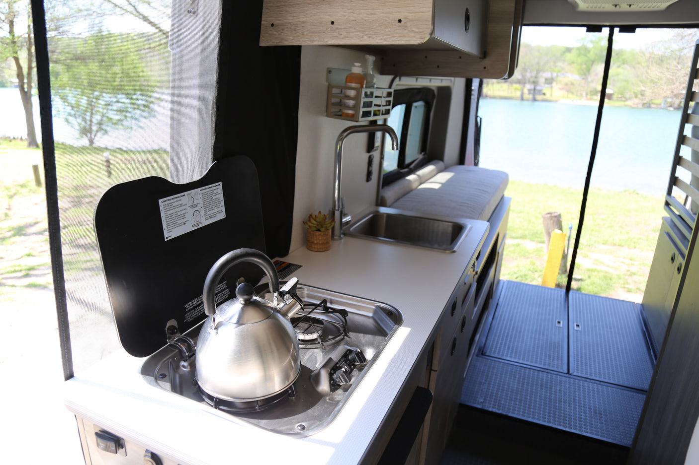 Enjoy your rv rental! Compact camper van kitchen with a stovetop, sink, and kettle, overlooking a serene lake.