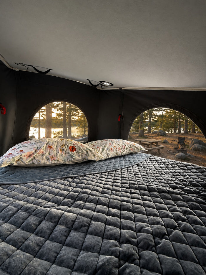 Cozy rv rental interior with a plush, quilted bed & floral pillows. Arched windows reveal a beautiful lake & forest view.