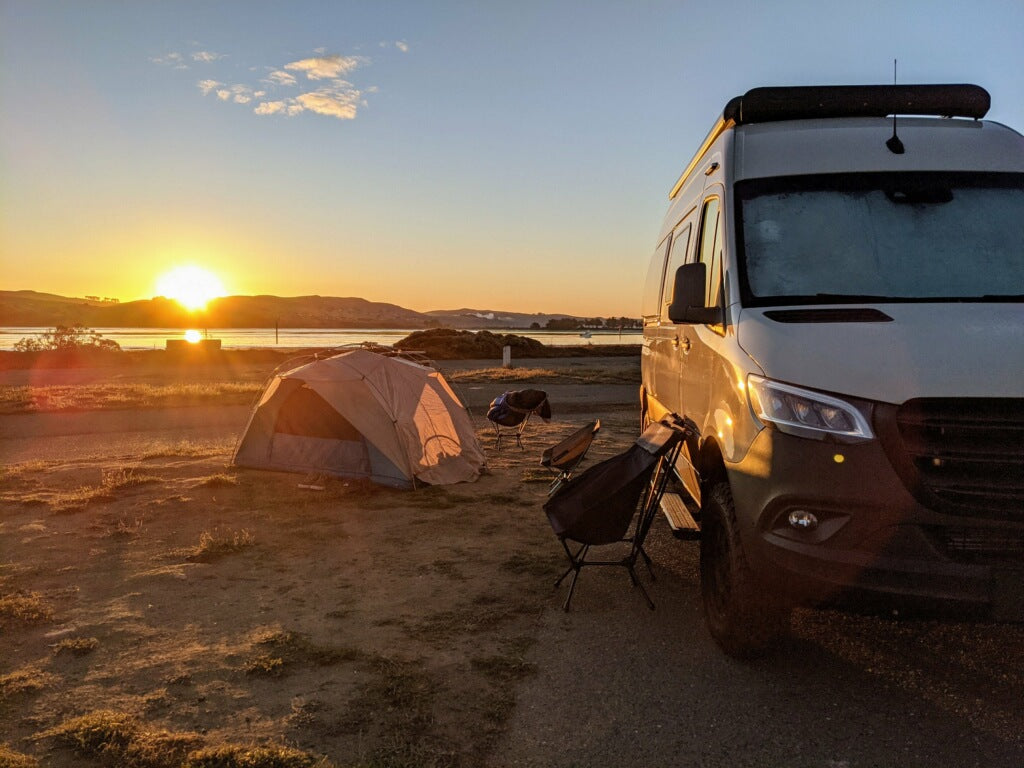 Stunning sunset camping scene with an RV rental, tent, and chairs by the water. Perfect outdoor adventure.