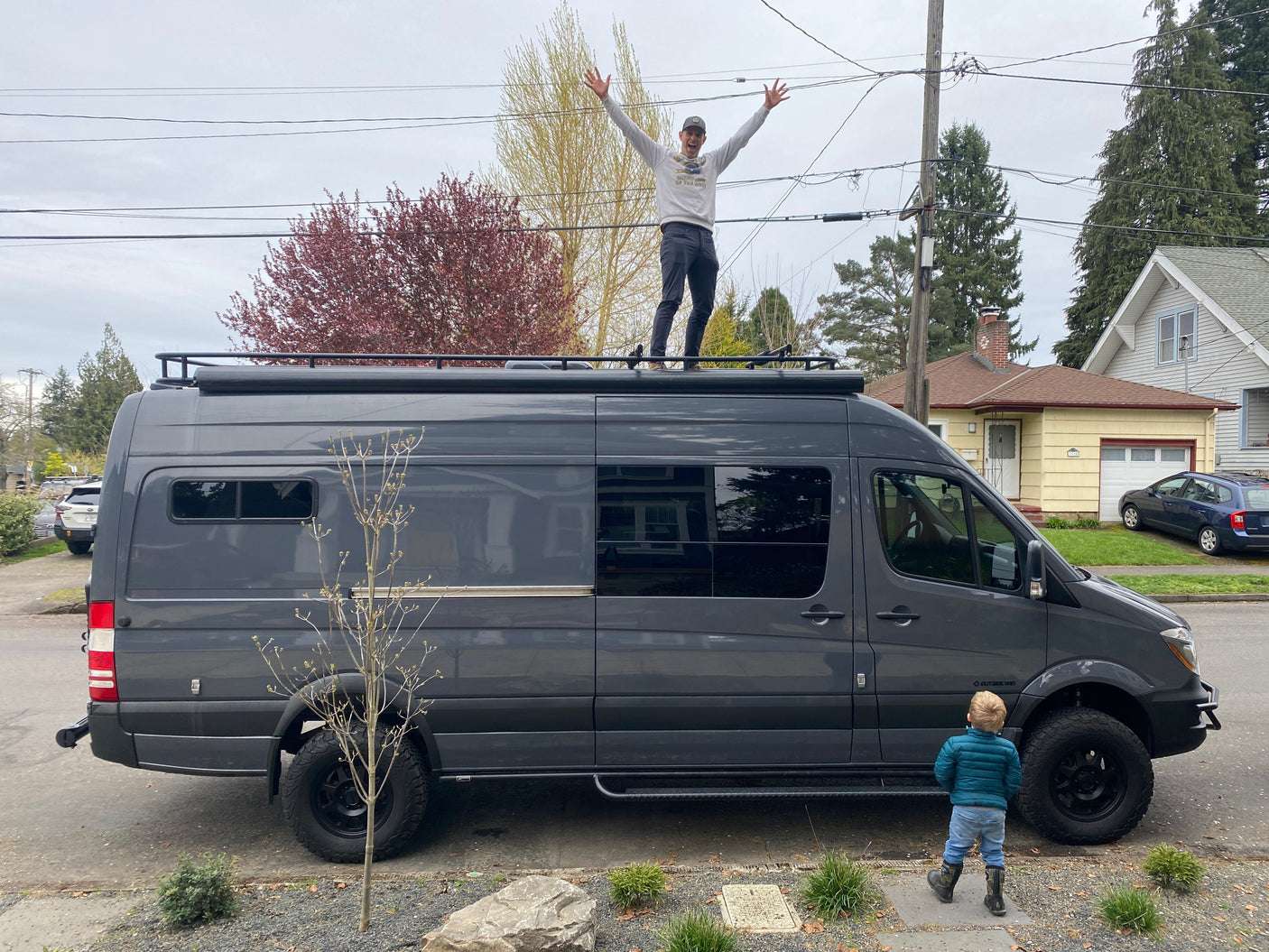 Man joyfully stands atop a sleek grey camper van (RV rental) with a child admiring it. Perfect for your next adventure!