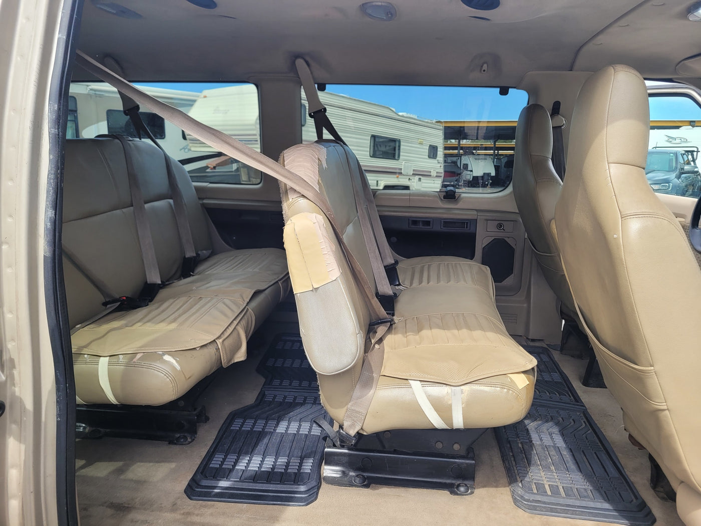 Interior of an **RV rental** van, showing two rows of tan seats with seatbelts, rubber floor mats, and parked RVs outside. — photo 8