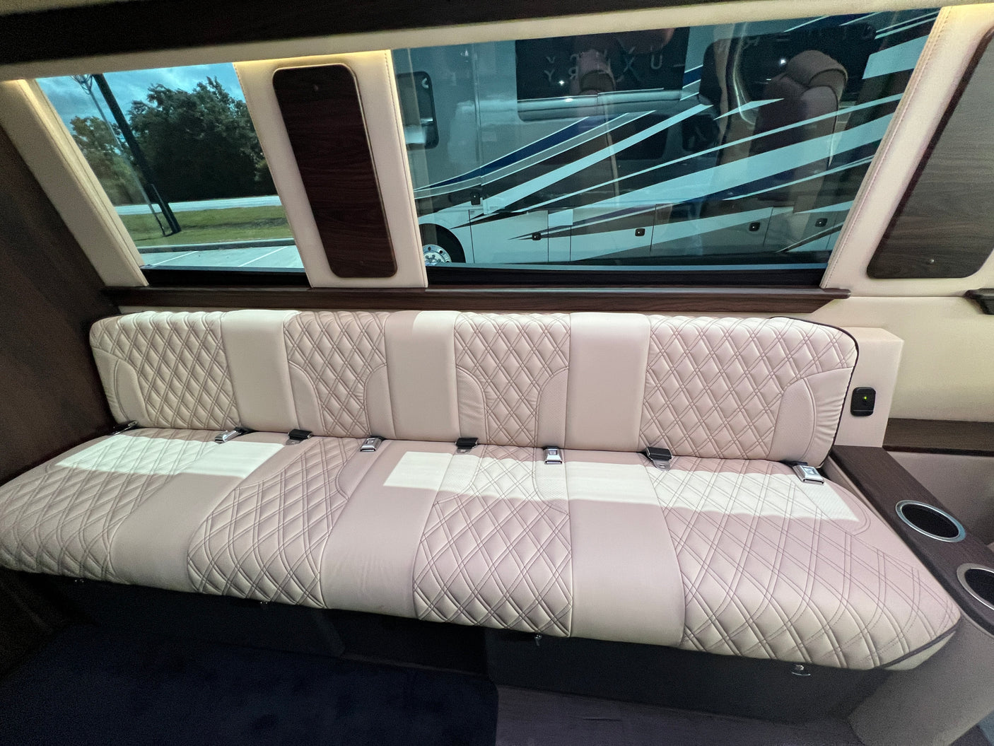 Inside a luxury RV rental: cream quilted leather bench seat with seatbelts, large windows reflecting another RV.