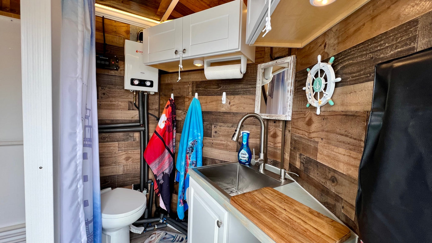 Compact RV rental interior featuring a functional kitchen sink, toilet, and efficient water heater in a wood-paneled space.