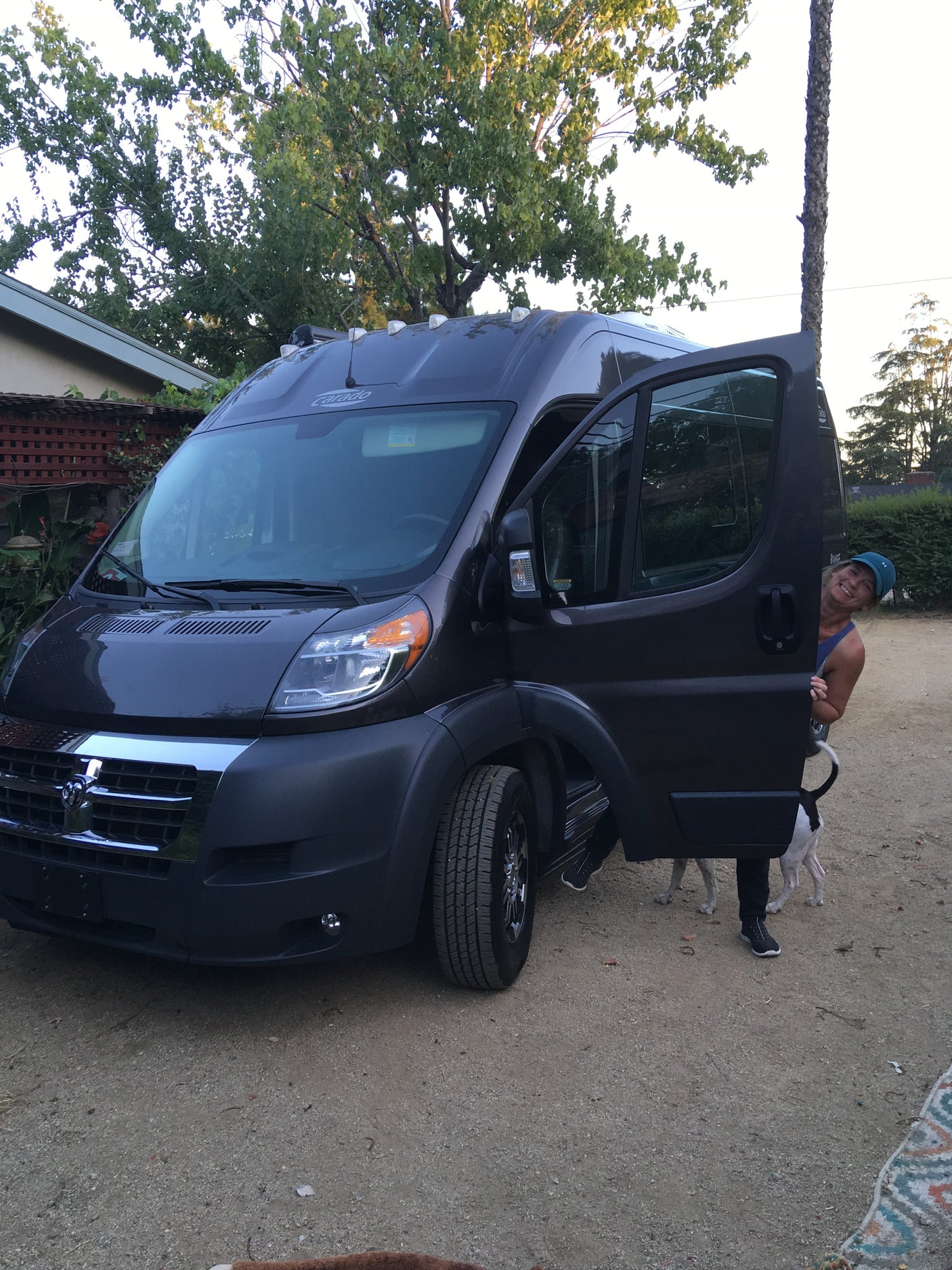 Smiling person and dog peeking from an open side door of a dark grey RV rental, parked on a dirt lot.
