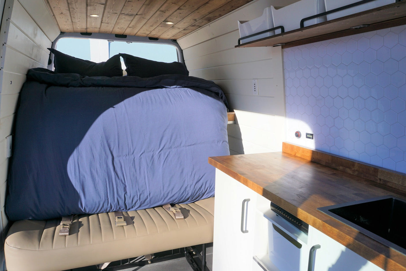 RV rental interior: Comfy bed over belted bench, wood counter, sink, and hex tile kitchen backsplash.