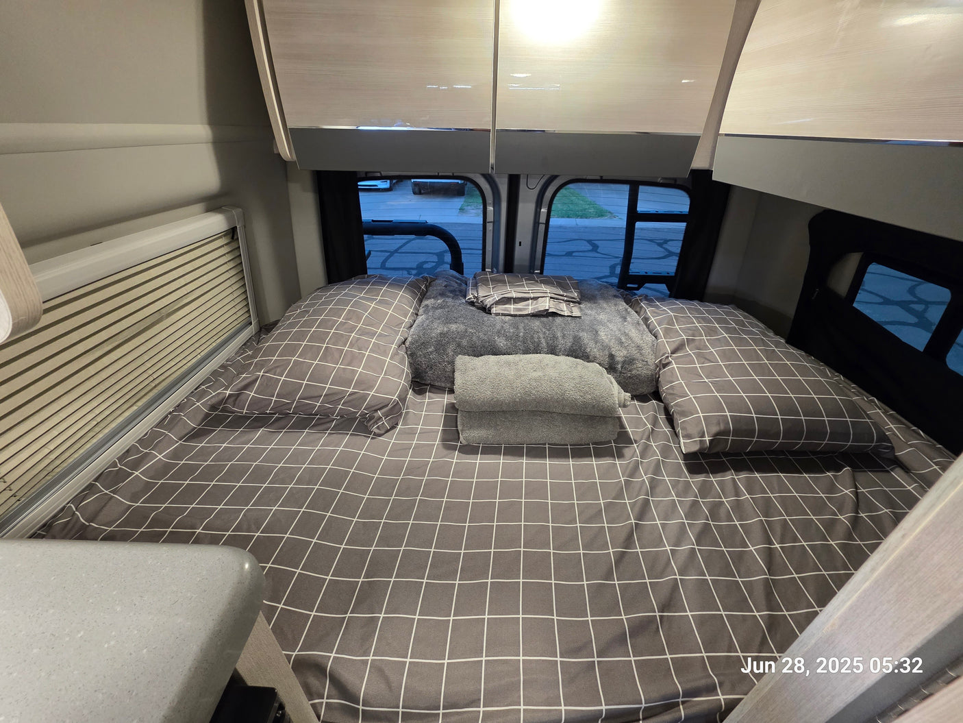 An RV rental bedroom features a cozy bed with checkered linens, practical overhead storage, and a view through rear windows.