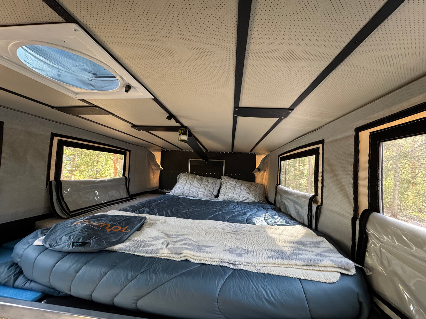 Inside a comfortable RV rental bedroom, featuring a large bed, side windows with tree views, and a Dometic ceiling vent.