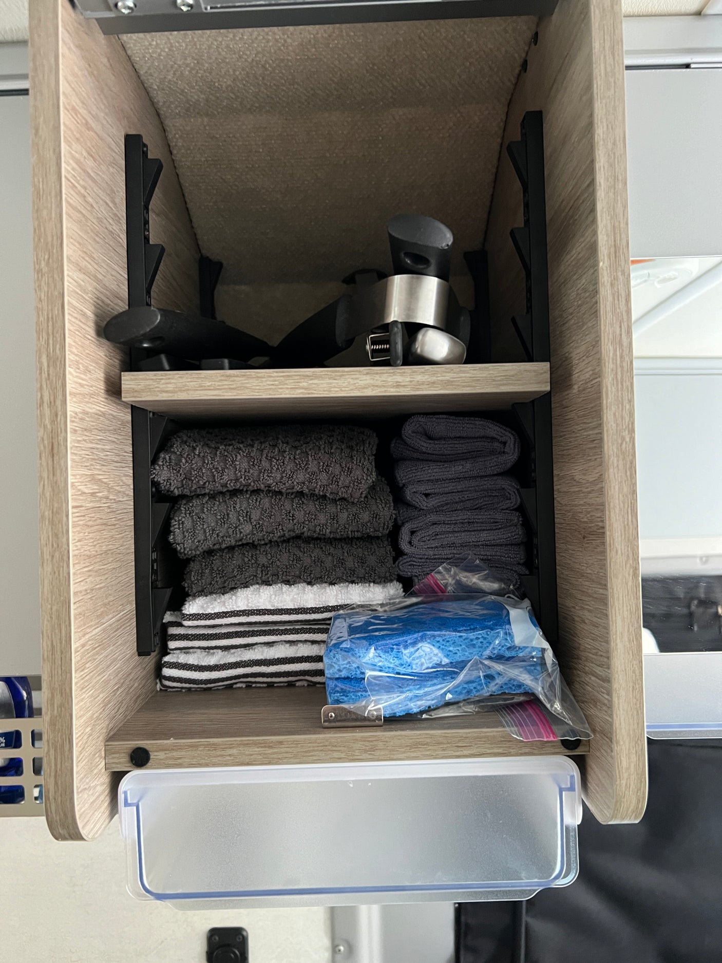 Organized RV rental cabinet features kitchen gadgets, various folded towels, and cleaning sponges, with a pull-out drawer.