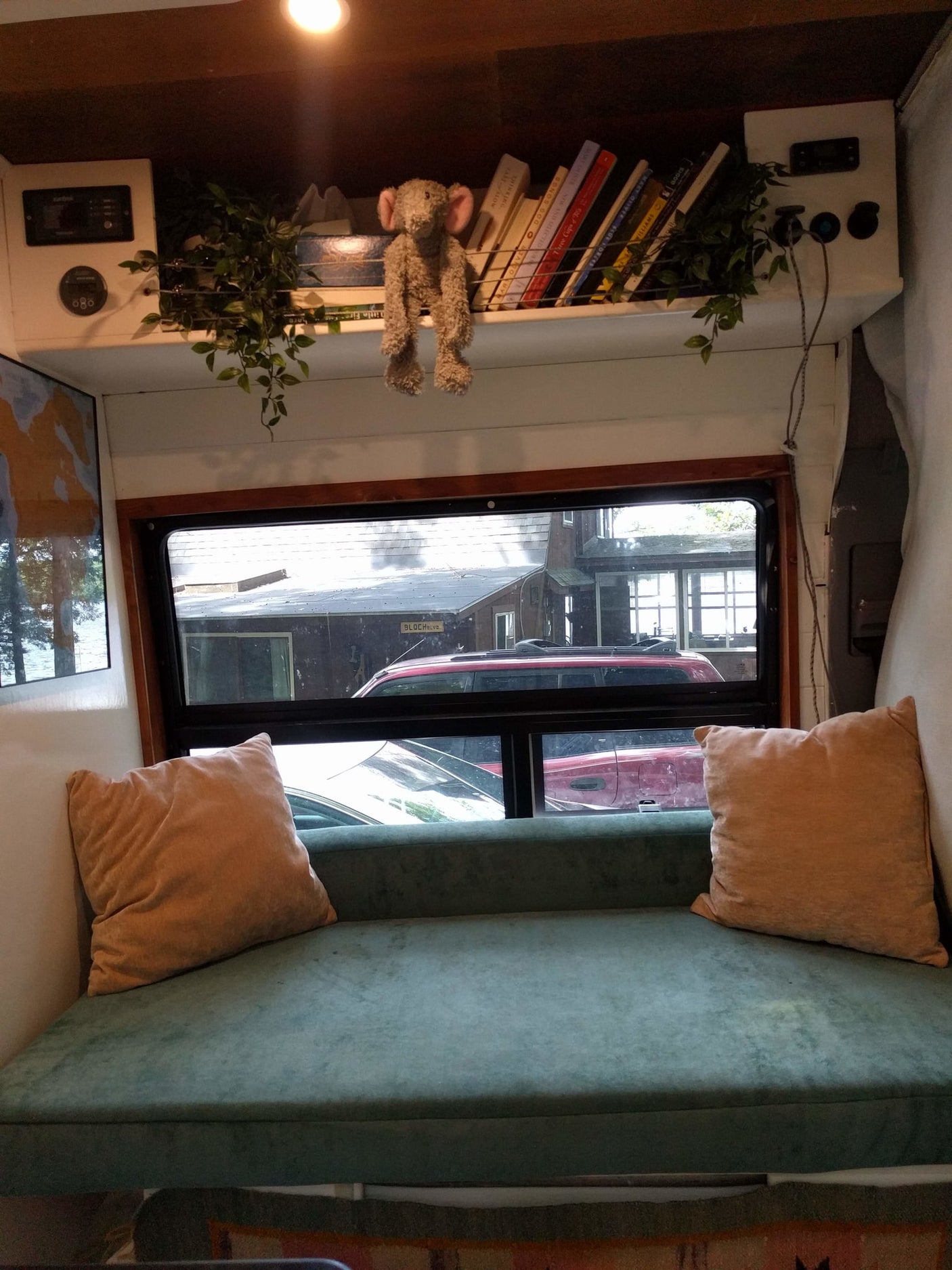 An RV rental's cozy interior features a teal cushion, pillows, bookshelf, controls, and a window with cars outside.