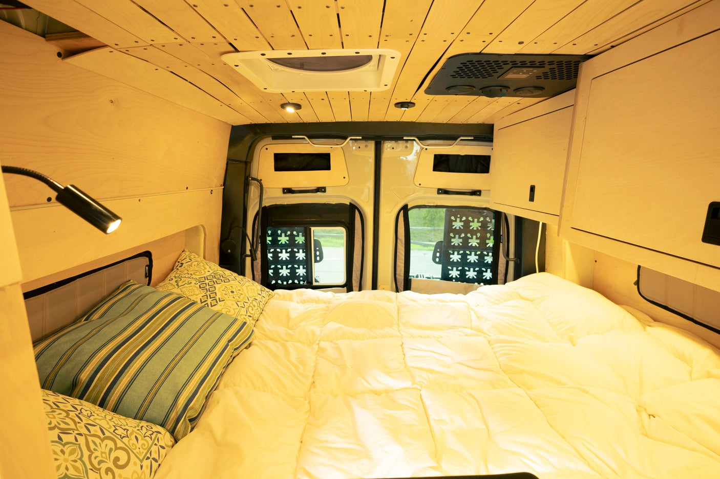 Inside a cozy RV rental: a bed with soft bedding, warm wood paneling, overhead storage, and ceiling climate units.