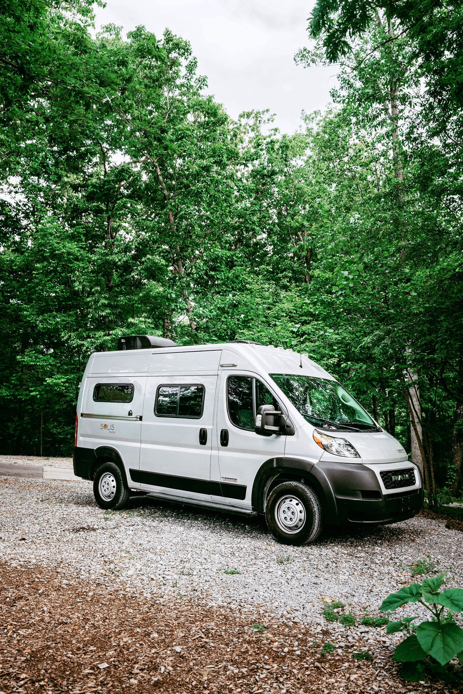 surrounded by lush green forest. Perfect for nature travel.|Winnebago Solis Pocket RV rental parked on gravel in a green