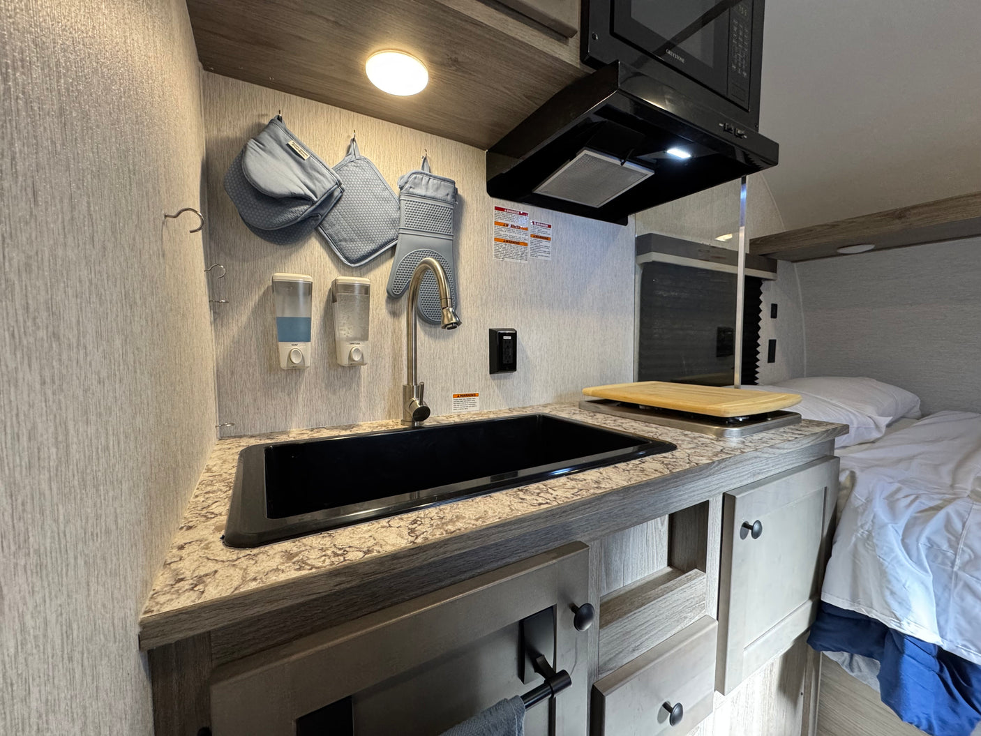 This RV rental features a compact kitchen with a black sink, microwave, and range hood, plus an adjacent bed for cozy travel.