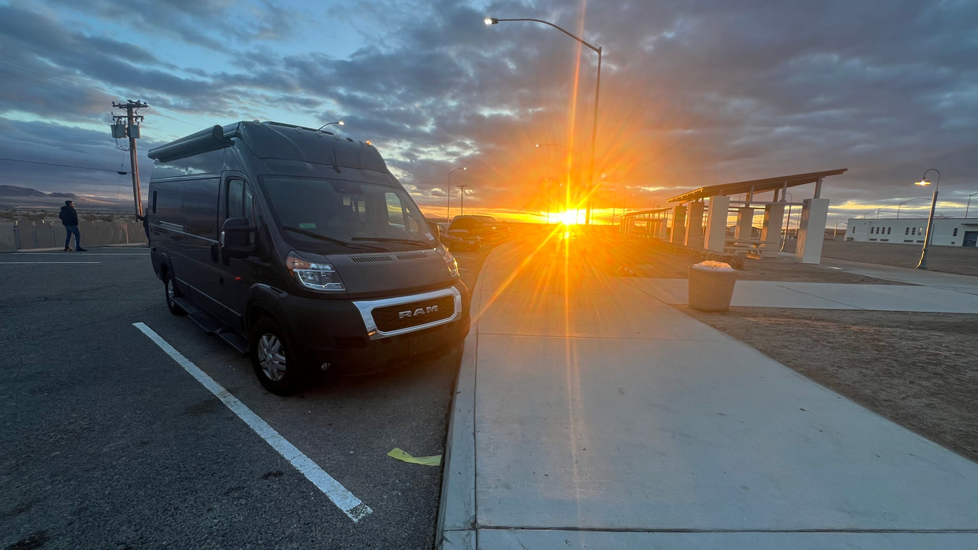 Dark gray RV rental van parked at a desert rest stop during a brilliant sunset, with a person, shelters, and bright sun.
