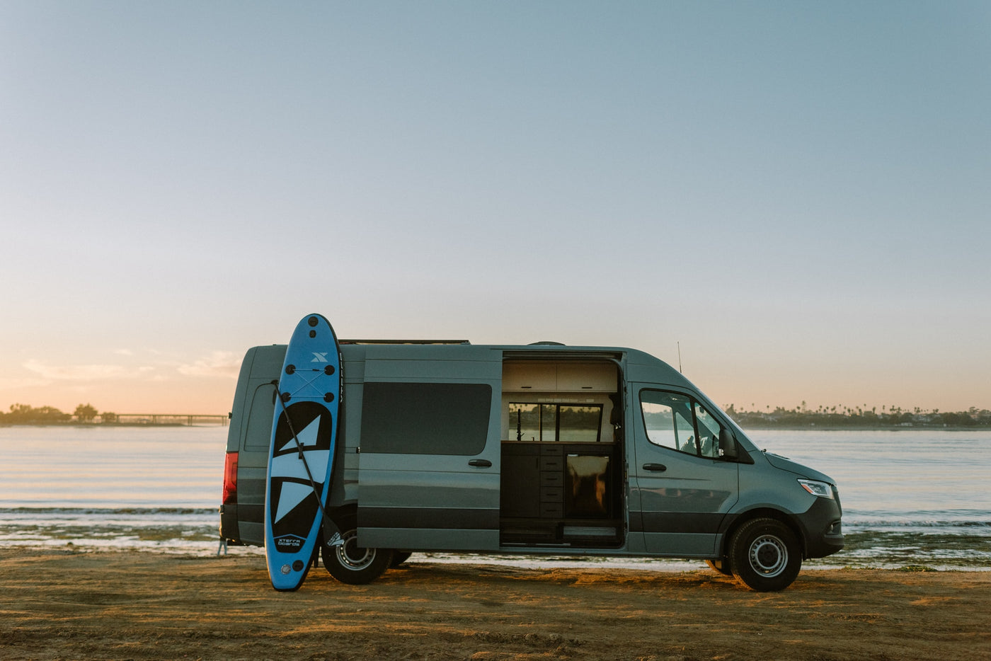 RV rental: Modern gray camper van on sandy beach at sunset. Open side door reveals custom interior. Blue paddleboard leans on rear. — photo 37
